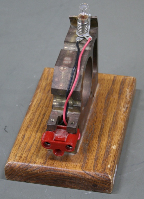 A flashlight lamp in a screw mount connected to banana socket terminals, supported on a block of scrap metal mounted on a wooden base.