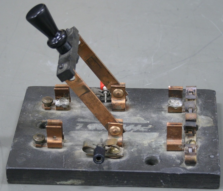 A double-pole knife switch mounted centrally on a weathered rectangular base throws to contacts on either side.  The switch has banana plug connectors and the contacts on one side connect to clips for holding wires.