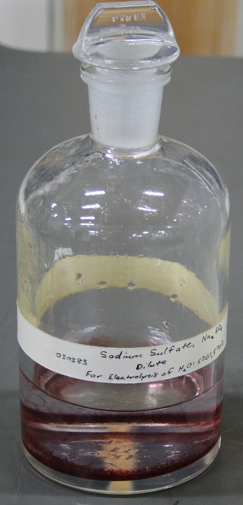 A glass flask about 25% full of clear liquid with solid precipitate around the bottom edge.  The label reads "sodium sulfate dilute for electrolysis of H2O".