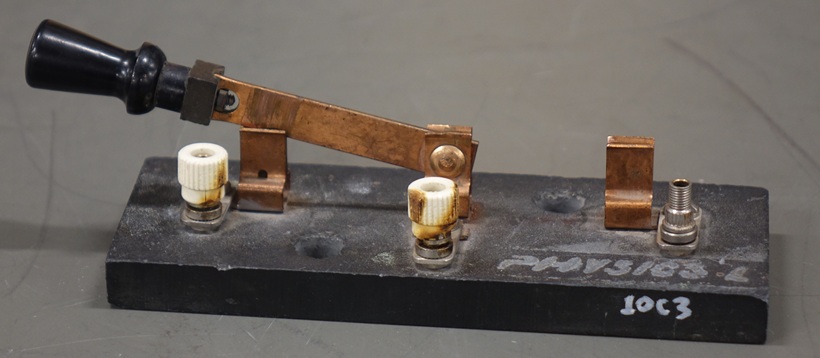 A single-pole knife switch mounted centrally on a weathered rectangular base throws to contacts on either side.  All three contact points have a banana plug connector.