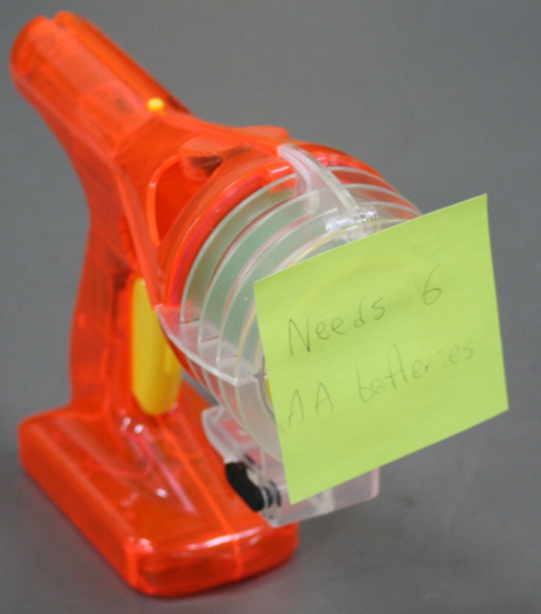 A bright orange plastic toy gun resembling a pulp science fiction blaster, with a reservoir for fog fluid.  A sticky note over the opening reads "Needs 6 AA batteries".