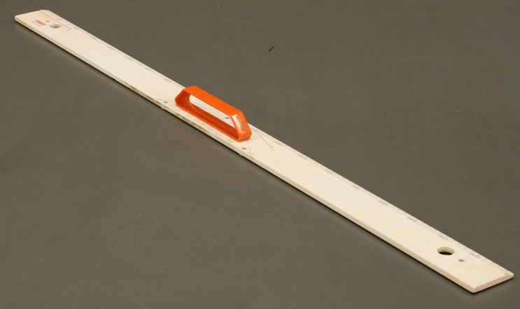 A two-inch wide plastic meter stick marked in centimeters.  Faded angle marks radiate from the midpoint every 15 degrees.  A handle is mounted in the center across the angle marks.