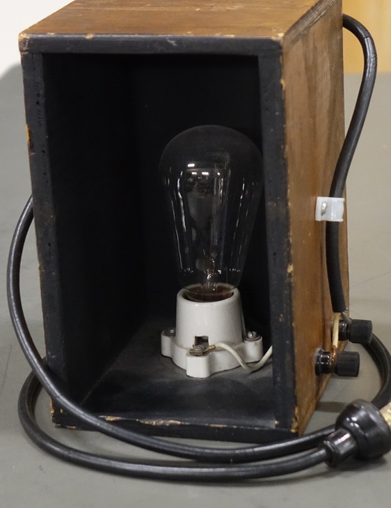 An incandescent light bulb with a looping filament, housed in a wooden box with an attached power cord.