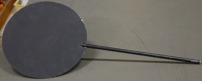 A head-sized circular sheet of metal, painted matte black, is mounted on a foot-long metal rod.