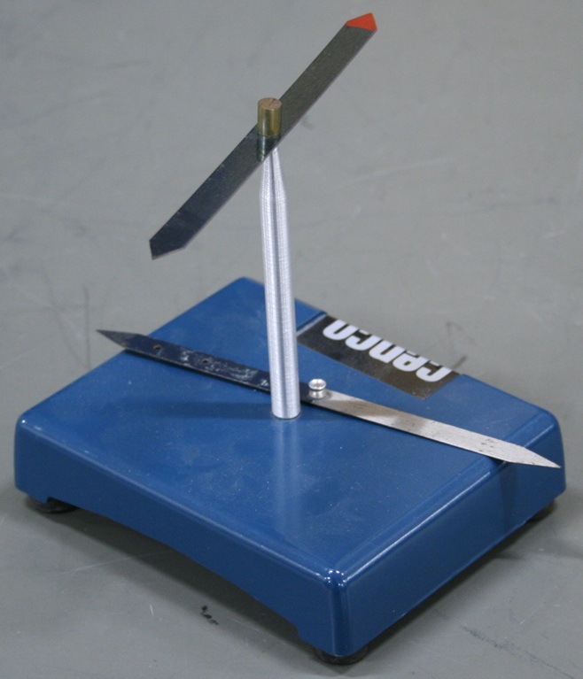 A short medal rod with a needle tip mounted on a rectangular base.  One compass needle is balanced on the tip, and another rests on the base.