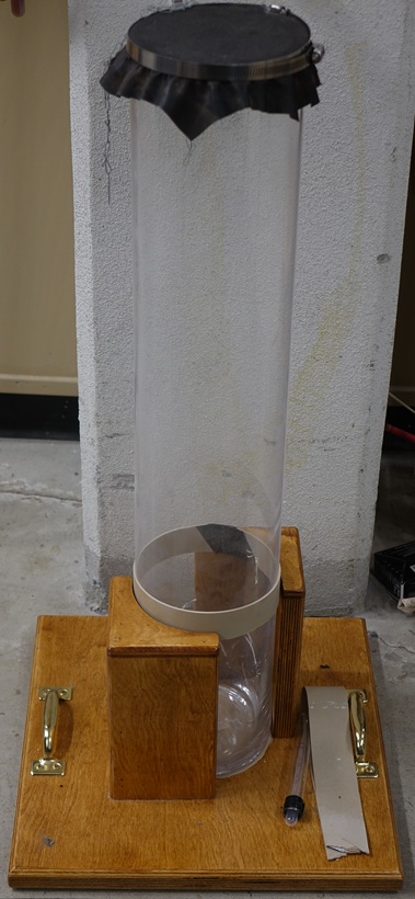 A cylindrical glass tank is mounted cetically on a wooden base with handles.  A hose clamp secures a sheet of rubber over the tank's opening.