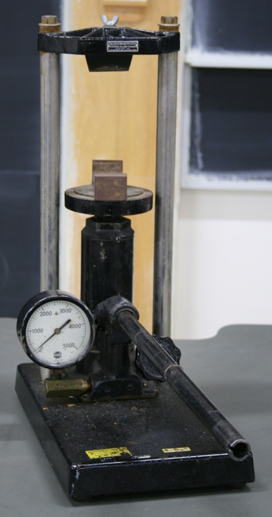 A hydraulic jack with a pressure gauge, manual lever, and release valve mounted on a heavy metal base.  A plate with two parallel upright bars 4.5 inches apart rests on the piston, which can be raised against a wedge suspended above the piston plate.