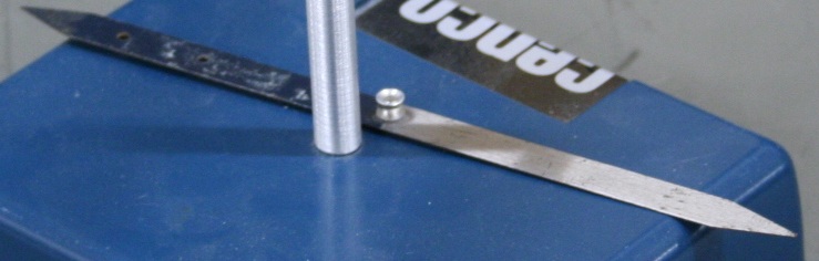 A compass needle six inches long with a central pivot mount.  North is painted dark blue.