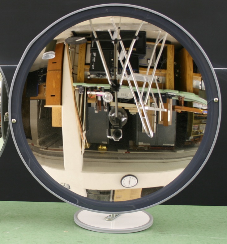 A plastic-backed concave mirror two feet across stands on a circular base and reflects an upside-down image of the lab it's facing.