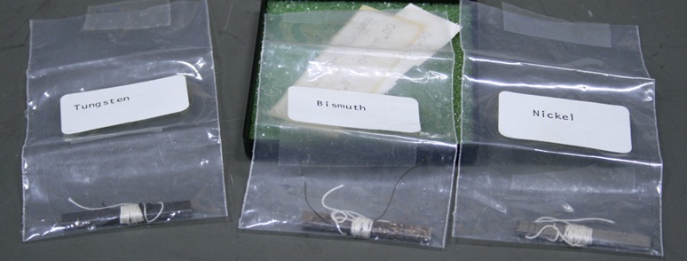 Three thin bar samples of tungsten, bismuth, and nickel, each an inch and a half long and in a tiny palstic bag.