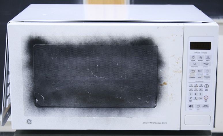 A stained and damaged microwave oven with its outer case loose.  The glass front has been removed and the grid painted black.