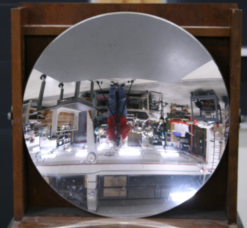 A round concave mirror a foot and a half across mounted in a wooden box frame reflects an upside-down image of the photographer and surrounding lab scene.
