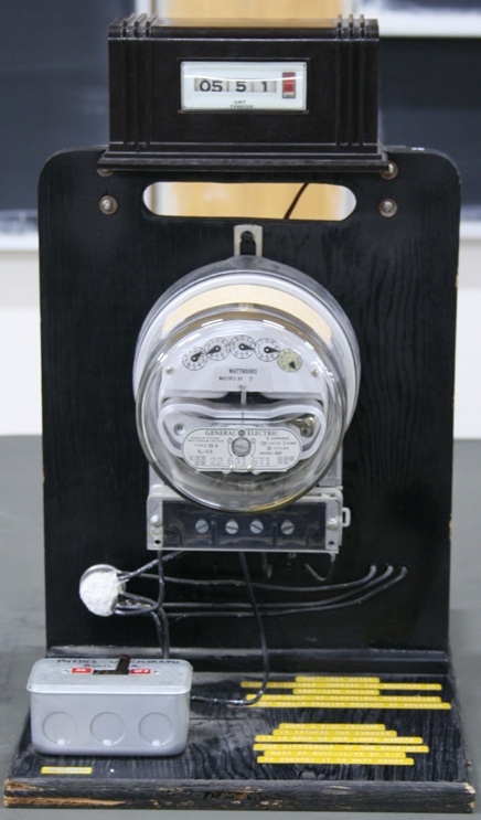 An old-fashioned dial power meter mounted on a wooden base, with a rolling timer above and a power switch below.