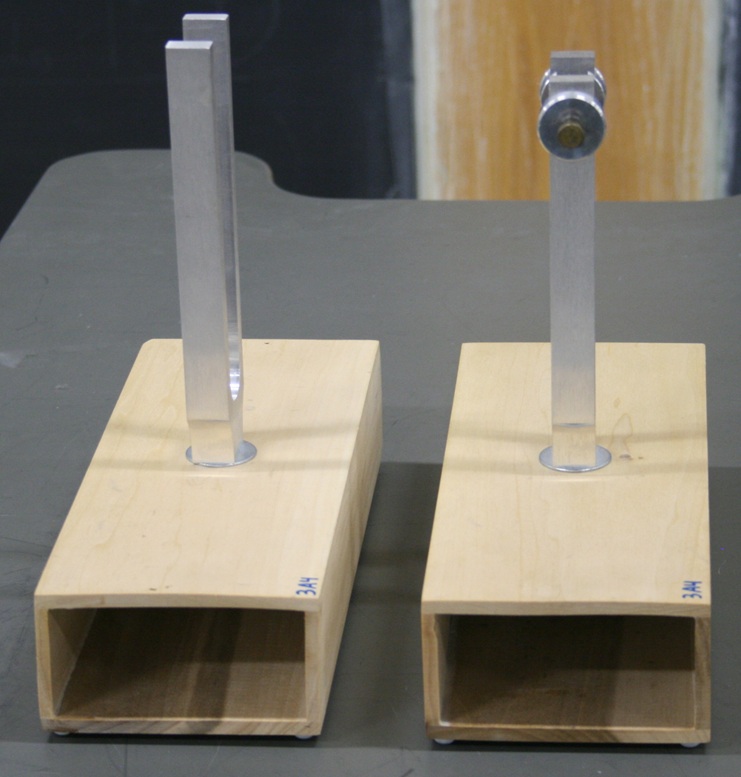 Two hollow wood boxes sit adjacent with the open ends facing forward.  Two rectangular-prong tuning forks are centrally mounted in the top faces of the boxes.  One of the forks has adjustable clamp masses to tune its pitch.