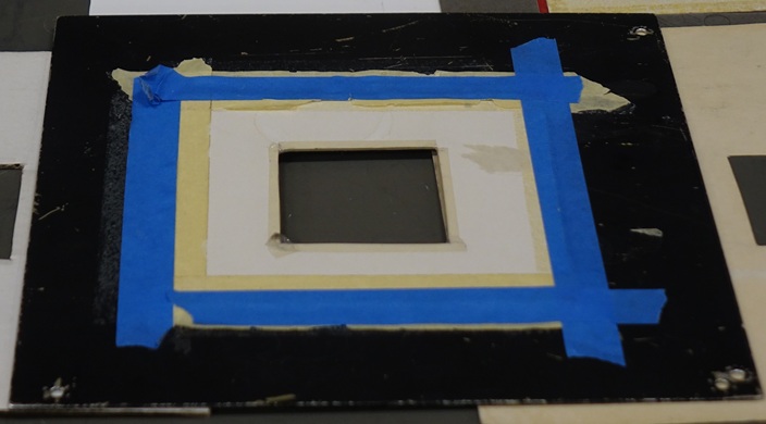 A square foot metal sheet with a 3-inch square mask taped over the center opening.