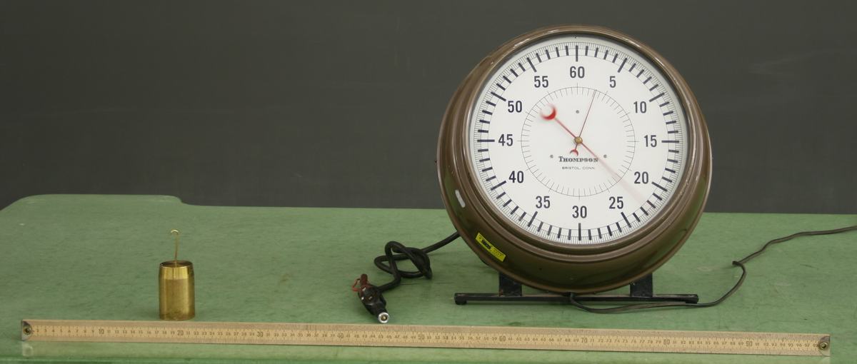 Brass cylindrical 1 kg mass, wooden meter stick, and large Thompson timer with 60-second dial displayed on a green demonstration table