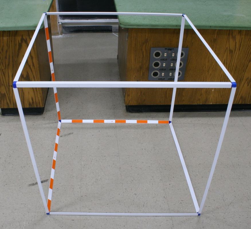 White tubular frame forming a one-meter cube, with three bottom edges marked by orange-white striped decimeter intervals and blue tape at the corners