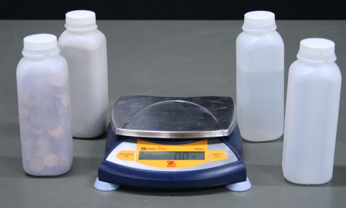 Four identical 473 mL translucent plastic bottles filled with different materials — one with pennies, one with white powder — arranged around an Ohaus Scout Pro 4000g digital scale