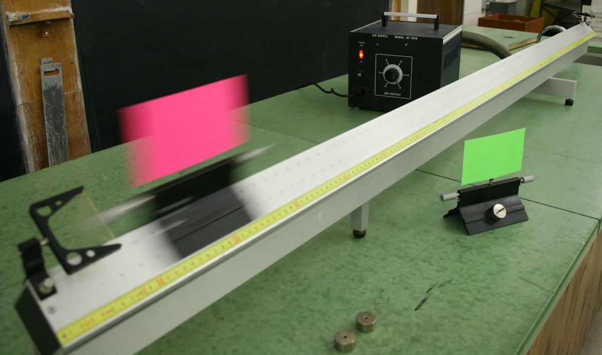 Closer view showing the black air blower unit with control dial behind the track, pink-flag glider in motion (blurred), green-flag glider stationary