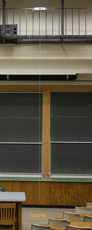 View from lecture hall floor looking up at the catwalk railing with the release platform mounted above, a string hanging down vertically toward the floor, chalkboards visible behind