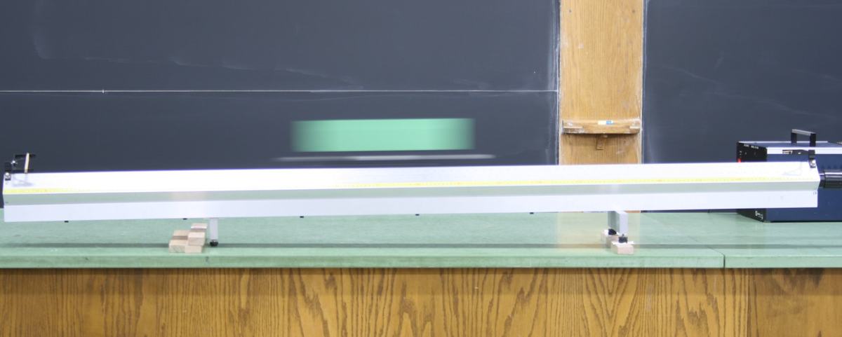 PASCO aluminum air track on a green lab bench with adjustable metal leveling feet, a yellow measurement scale along the track, wooden blocks underneath for tilting, and the air blower unit at the right end