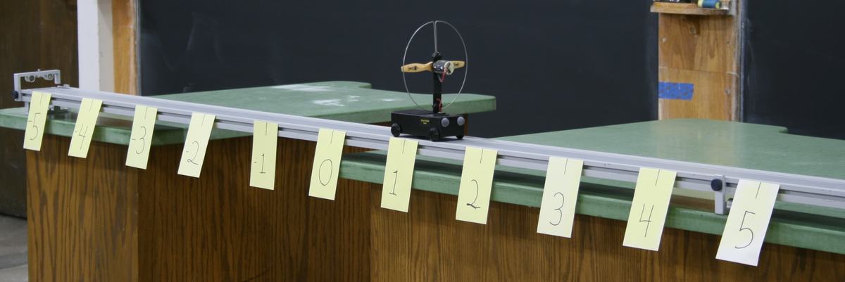 PASCO fan cart with wooden propeller and guard ring on a black base, sitting on an aluminum dynamics track spanning two green demo tables, with yellow sticky-note markers numbered -5 to 5 hung at equal intervals along the track