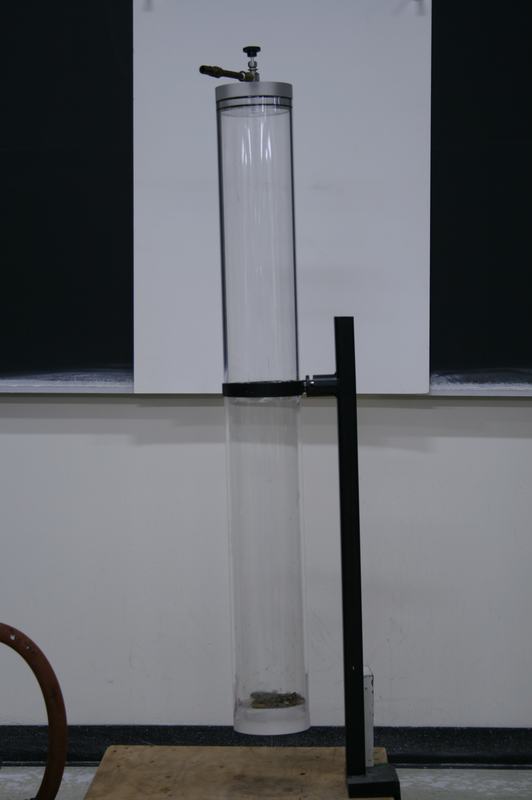 Clear acrylic cylinder mounted horizontally on a black metal bracket attached to a bench support, with a valve fitting at one end for vacuum pump connection