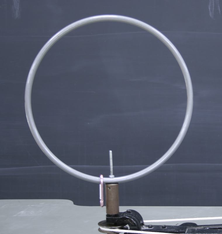Metal hoop mounted vertically on a brass spindle attached to a black hand-cranked centrifugal machine, with a small carabiner hanging at the bottom of the hoop