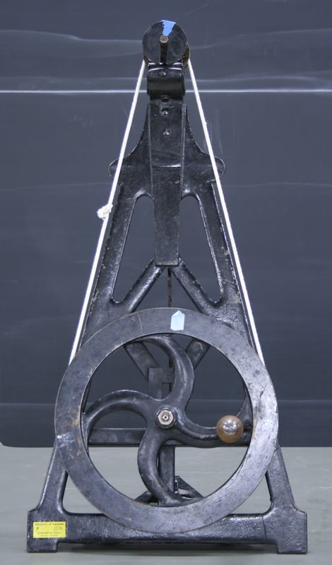 Cast-iron A-frame centrifugal machine with large hand-crank wheel at bottom, belt drive to small pulley at top of the frame, yellow warning label on base