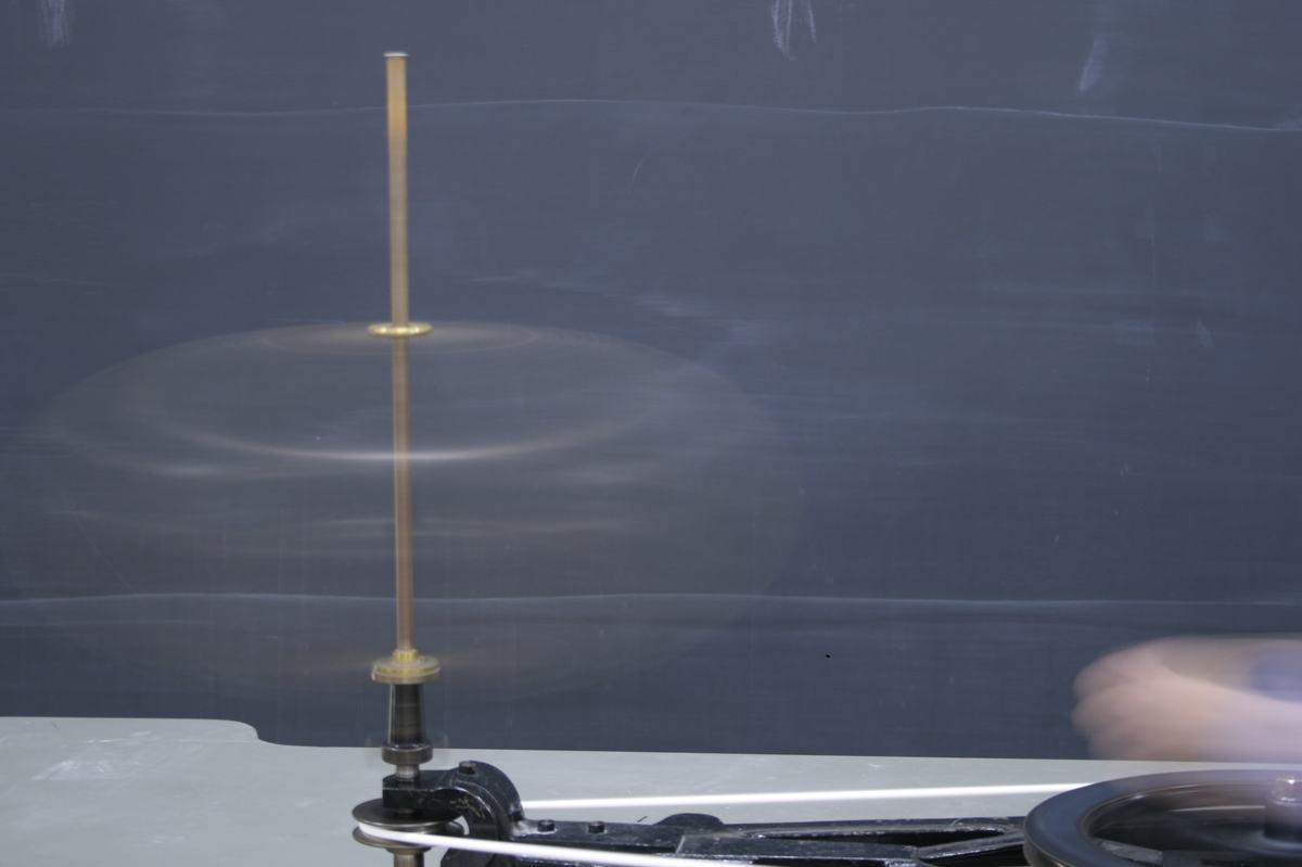 Flexible metal hoop spinning on a brass rod mounted on a hand-cranked centrifugal machine, showing oblate deformation with motion blur