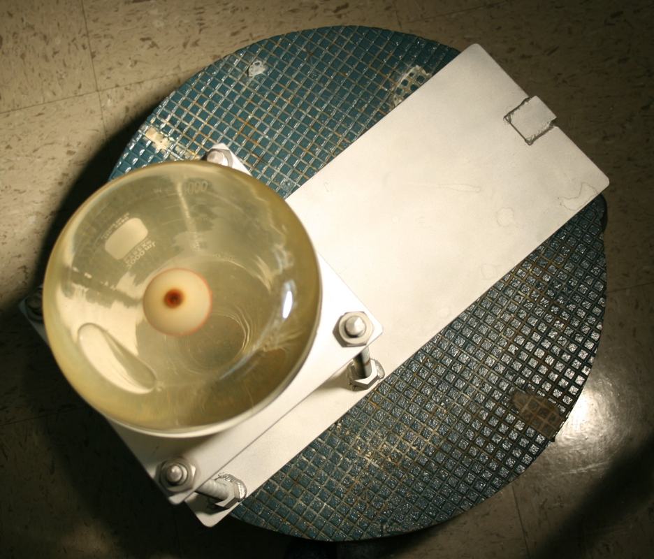 Side-angle overhead view of inverted Erlenmeyer flask with orange fishing float in white aluminum frame on teal rotating platform with grid markings