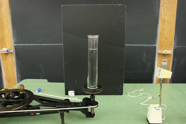 Glass cylinder mounted on a centrifugal machine with hand crank and belt drive, positioned in front of a black backdrop, with a desk lamp to the right