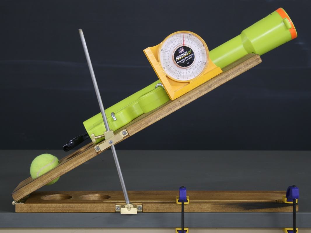 Spring-loaded tennis ball launcher: bright green tube with orange tip mounted on a wooden platform, Johnson angle locator attached, metal support rod setting the angle, tennis ball at the base, platform clamped to bench with two clamps