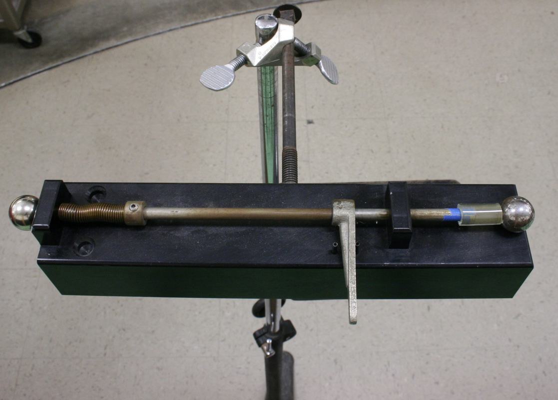 Free fall apparatus with a horizontal brass rod holding two steel balls on a green and black base, spring-loaded launch mechanism on one end, vertical trigger lever hanging below