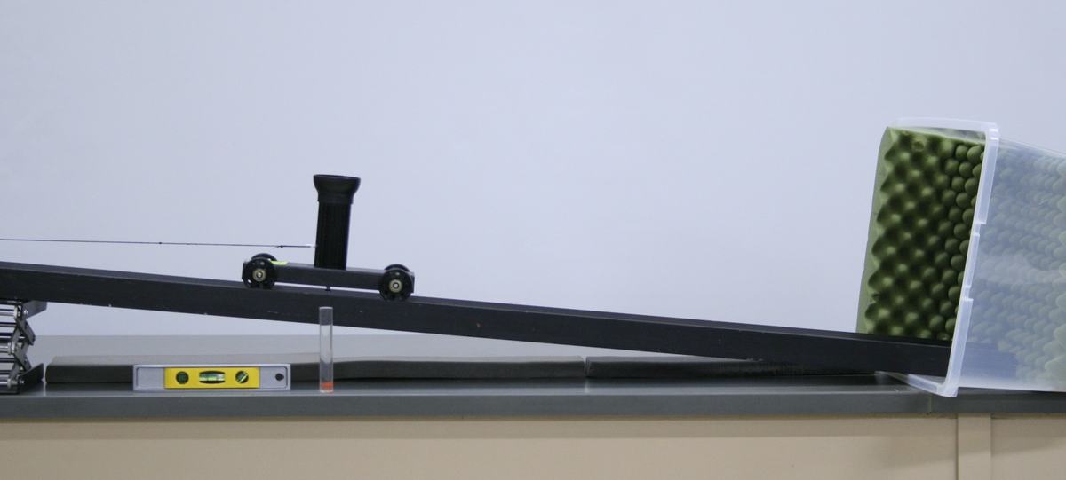 PASCO cannon cart on an inclined black track with vertical launch barrel, spirit level below the track, lower end nesting into a clear plastic bin lined with green eggshell foam