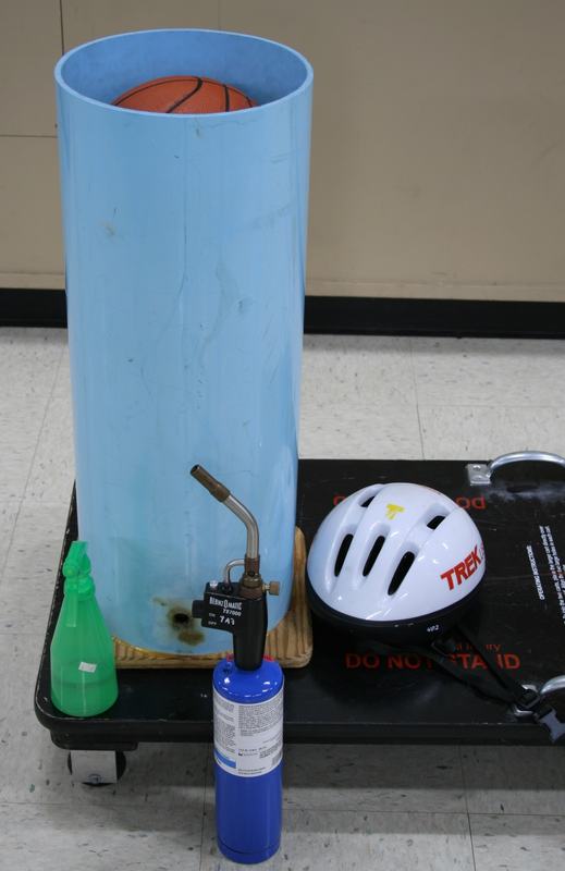 Light blue PVC cannon tube with basketball seated in top, mounted on black physicist's skateboard, with green ethanol spray bottle, propane torch, and white Trek bicycle helmet