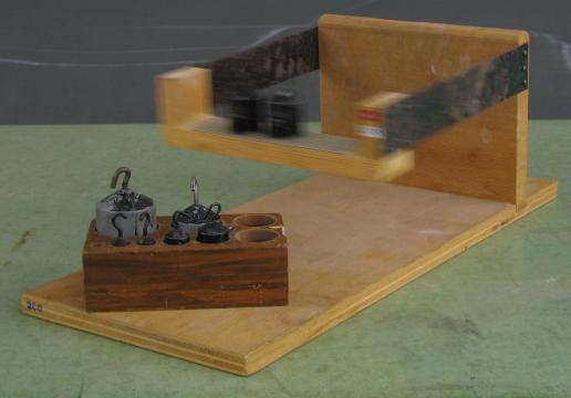 Inertia balance: a wooden base with two upright supports holding flexible metal blades that extend horizontally to a wooden oscillating platform, shown mid-oscillation as a blur. A calibrated mass set sits on the base beside it.