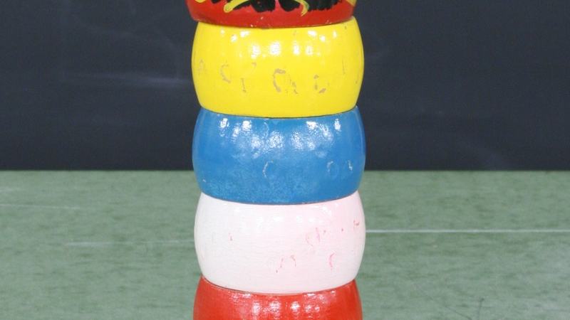 Daruma Otoshi toy: stacked colored wooden cylinders (green, white, red, pink, blue, yellow) with a painted daruma face on top and a small wooden hammer piece