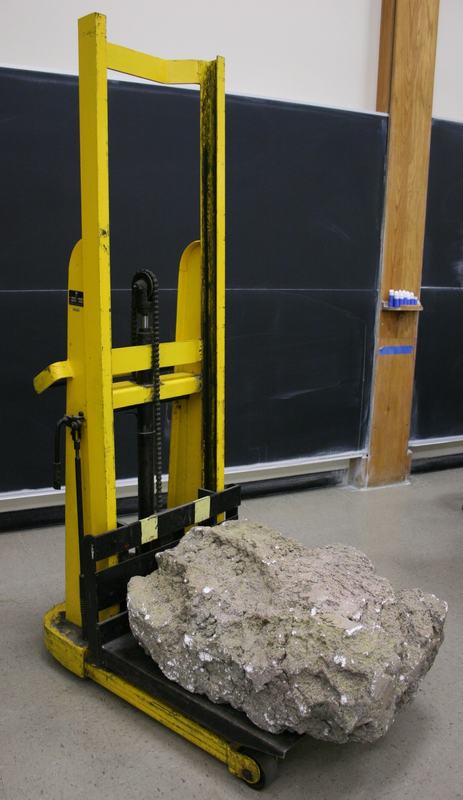 Large styrofoam boulder with realistic rough, weathered surface on the forks of a yellow hydraulic forklift in a classroom