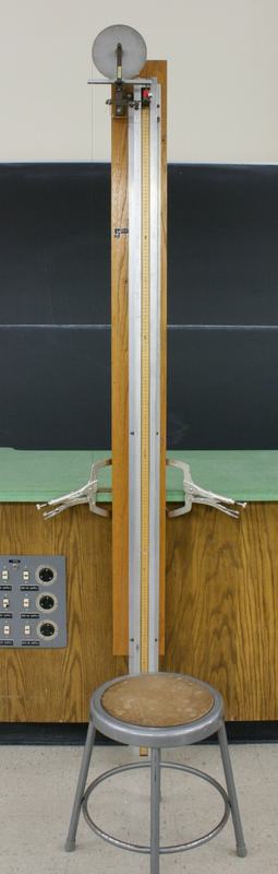 Tall wooden frame clamped to lecture bench with vise-grip clamps, metal disk pulley at top, meter stick mounted vertically on the frame, and a small metal stool at the base for height compensation