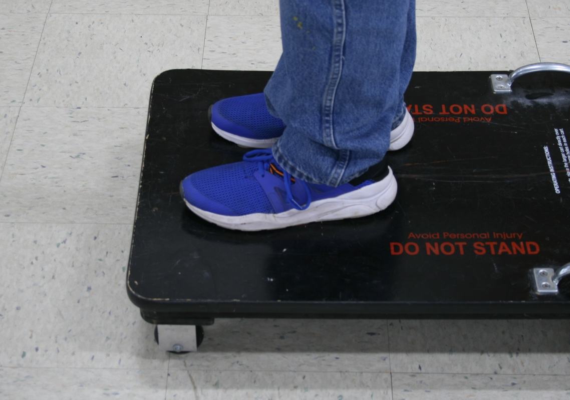 Person in blue sneakers and jeans standing on a large black wheeled platform with caster wheels on tile floor, red warning text reading 'Avoid Personal Injury DO NOT STAND' visible on the surface, metal handle at one end