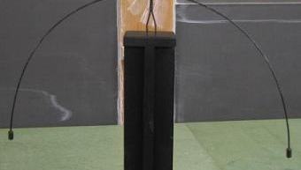 Wooden figure with round white head standing on a black pedestal; long curved wire arms arc downward on both sides with weights at the tips, lowering the center of mass below the feet