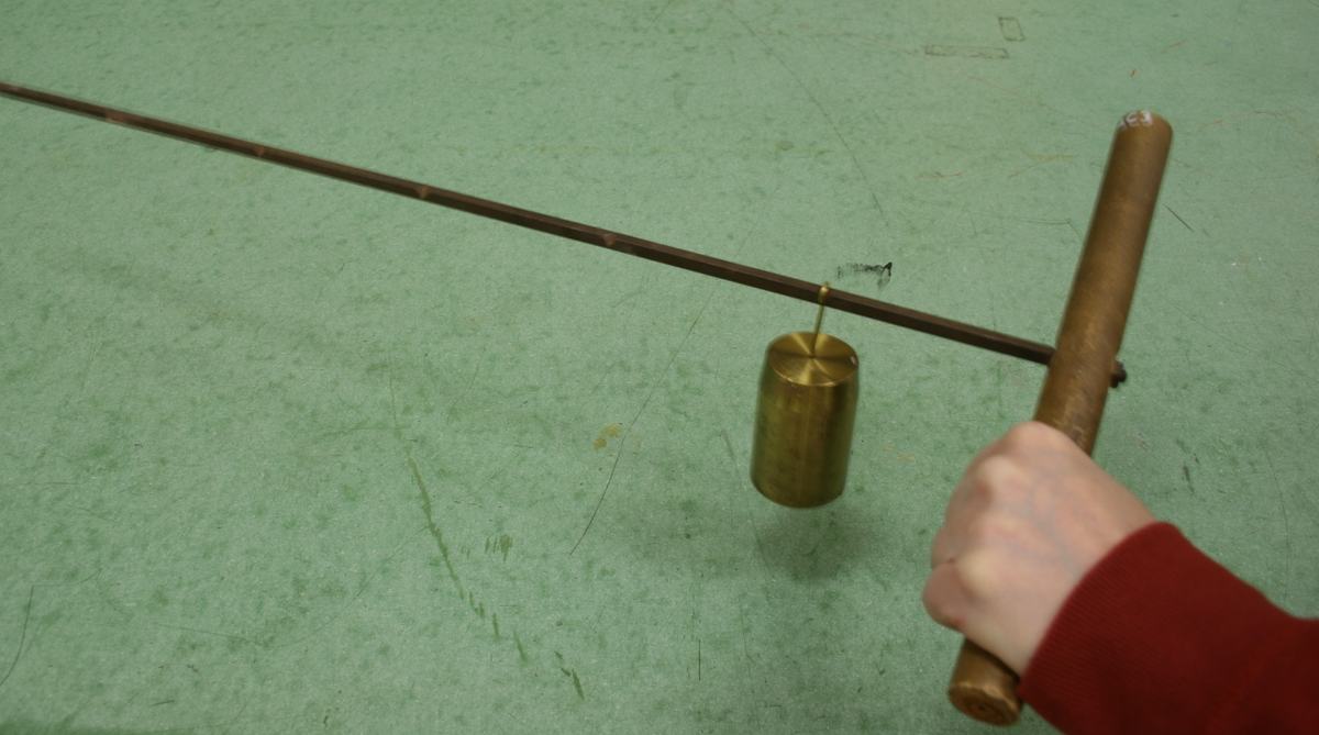 Hand gripping wooden cylindrical handle with thin metal rod extending left; a brass 1 kg mass hangs from a notch near the middle of the rod