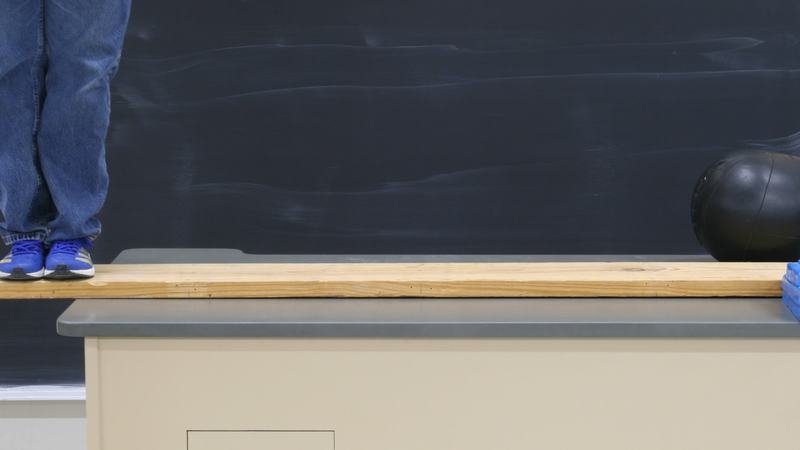 Person standing on the overhanging end of an 8-ft wooden plank on a lecture bench; medicine ball and blue safety-stop blocks visible at the far end, chalkboard behind
