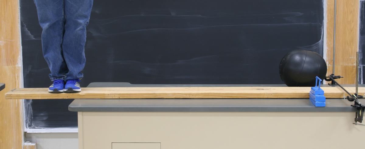 Person standing on the overhanging end of an 8-ft wooden plank on a lecture bench; medicine ball and blue safety-stop blocks visible at the far end, chalkboard behind