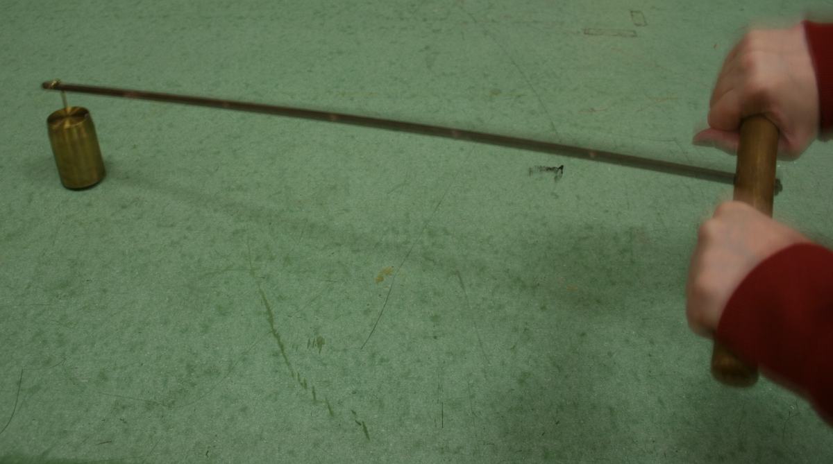 Two hands gripping the wooden handle as the brass mass hangs from a notch near the far end of the rod, demonstrating the increased difficulty