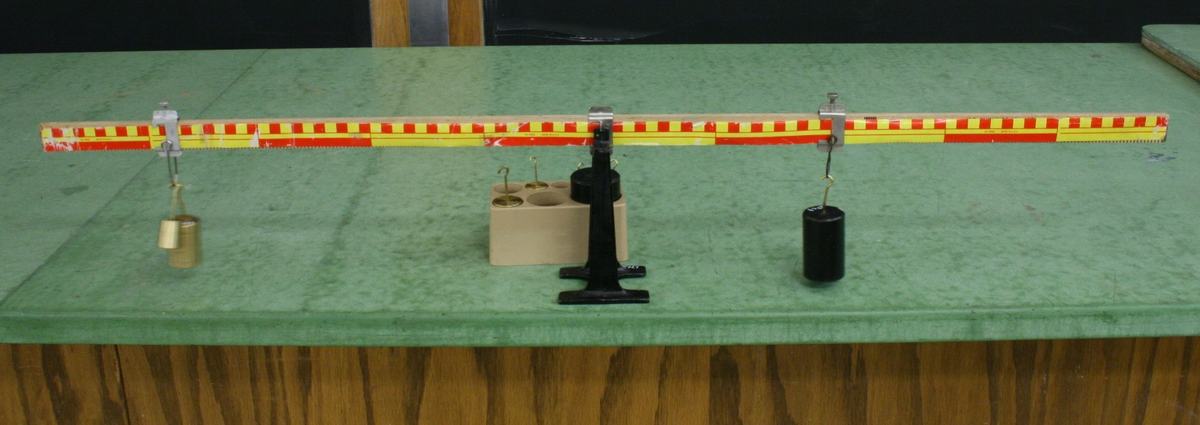 Red-and-yellow checkered meter stick balanced on a black pivot stand, with a brass mass hanging from a screw clamp on the left and a larger black cylindrical mass on the right, mass set visible behind the pivot