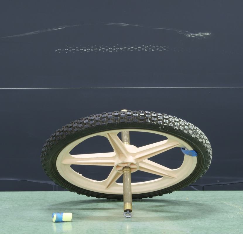 Wheel with tan plastic spokes and knobby black rubber tire on a metal axle, with chalk nearby and tread-pattern marks visible on the chalkboard behind