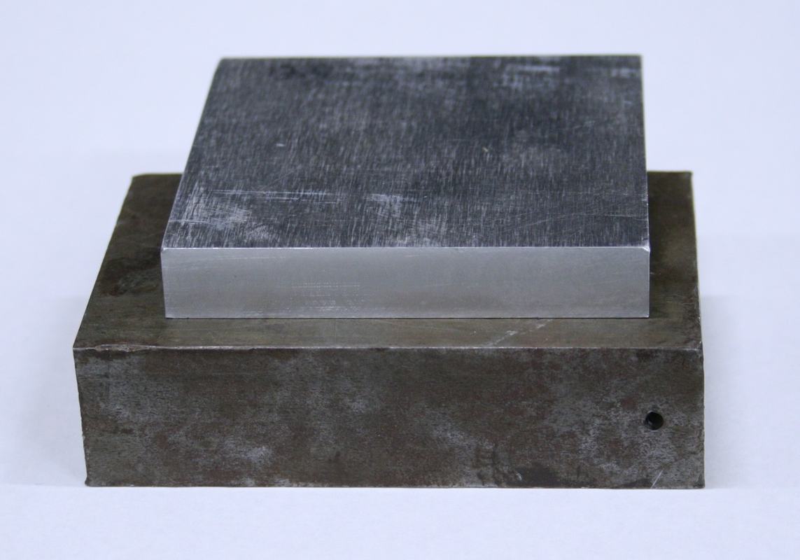 A smaller polished metal block resting on a larger darker metal block, both showing scratched and worn surfaces