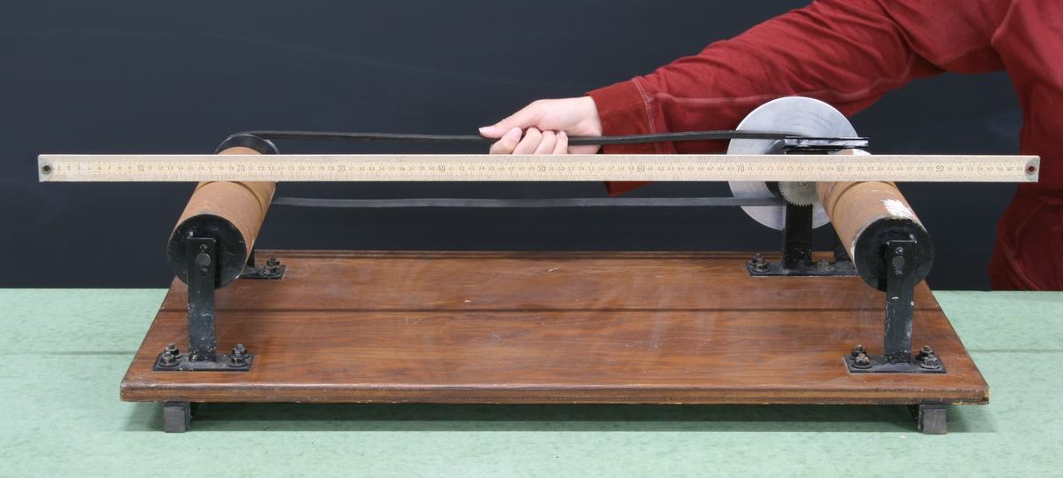 Two large tan cylindrical rollers mounted on black metal bearings on a wooden base, with a meter stick resting across them. A hand positions the stick. A pulley and belt are visible on the right side.